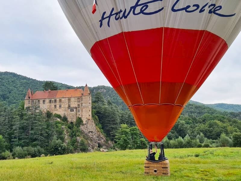 Montgolfière rouge et blanche survolant un château ancien entouré de verdure au Puy-en-Velay.