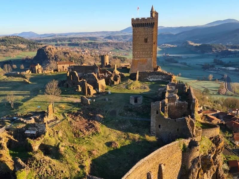 Vue aérienne d'un château médiéval en ruines avec une grande tour au centre sous un ciel clair.