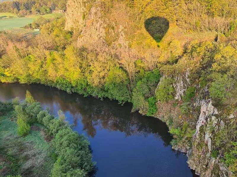Ombre d'une montgolfière projetée sur une falaise boisée au-dessus d'une rivière calme.