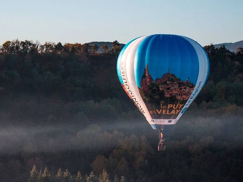 Montgolfière bleue avec image du Puy-en-Velay volant au-dessus d'une forêt au lever du jour.