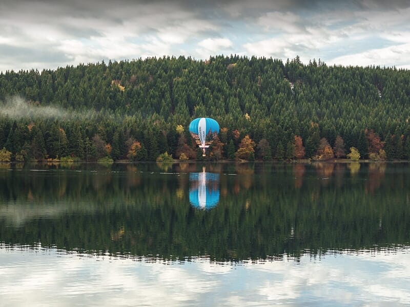 Montgolfière bleue survolant un lac calme avec forêt dense en arrière-plan sous lors d'un vol en montgolfière près de Langogne.