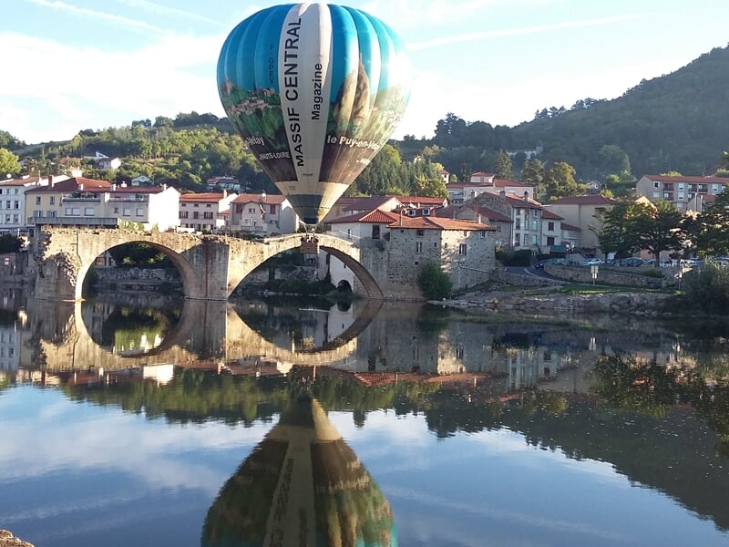 Montgolfière bleue reflétée dans l'eau calme d'une rivière avec un pont en pierre lors d'un vol en montgolfière près de Langogne.