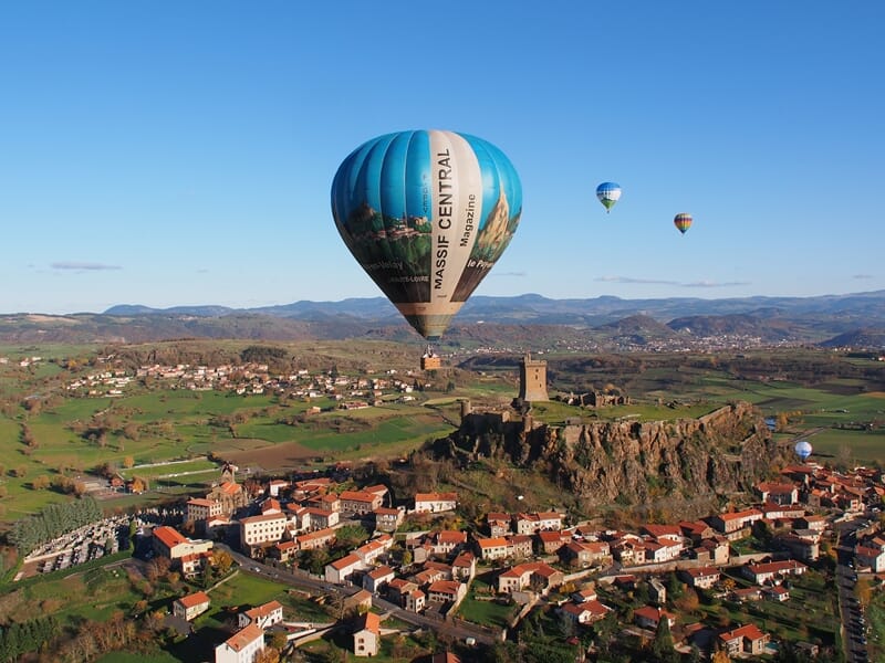 Montgolfière bleue survolant un village avec château sur une colline lors d'un vol en montgolfière près de Langogne.