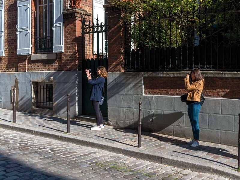 Deux personnes photographiant un portail en fer forgé dans une rue pavée ensoleillée à Paris.