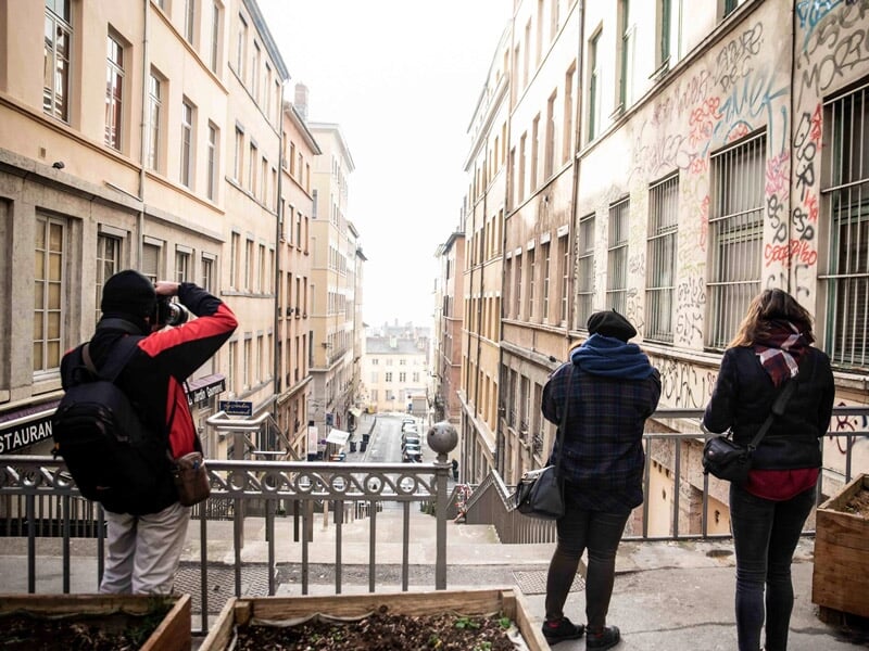 Trois personnes photographiant une rue en pente avec des bâtiments anciens sous un ciel clair à Lyon.