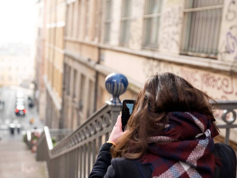 Femme prenant une photo avec un smartphone en haut d'un escalier urbain bordé de bâtiments anciens.