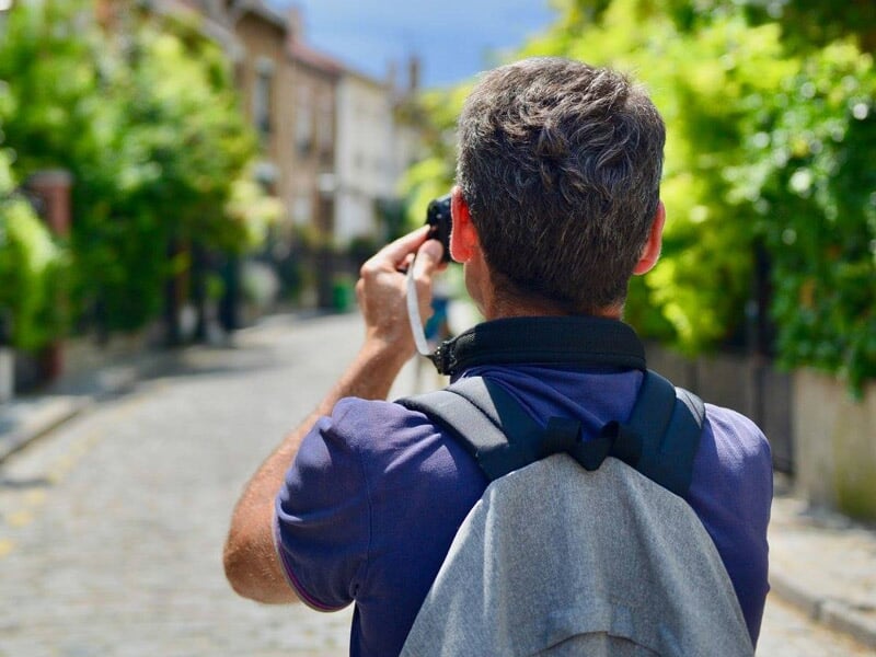 Homme avec sac à dos prenant une photo d'une rue pavée bordée d'arbres verts en plein jour.