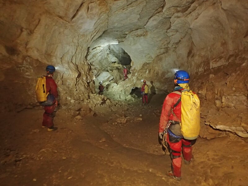 Explorateurs en combinaison rouge et casque bleu dans une grande grotte rocheuse éclairée par leurs lampes.
