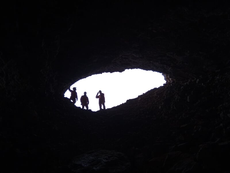 Silhouettes de spéléologues à l'entrée d'une grotte, contre un ciel lumineux et clair.