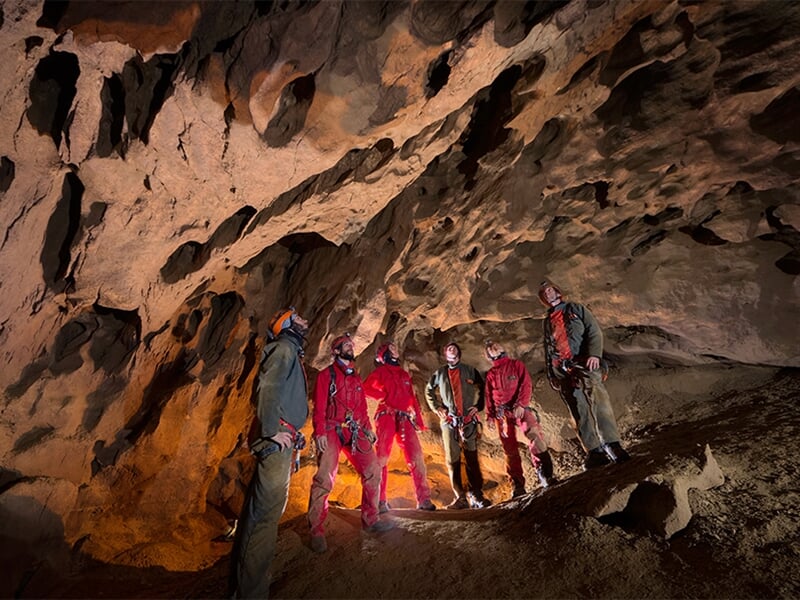 Groupe de spéléologues en combinaison rouge et casque dans une grande salle souterraine éclairée.