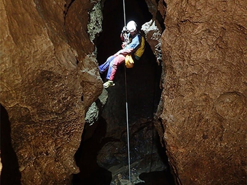 Spéléologue équipé descend en rappel dans une grotte étroite et sombre avec un casque lumineux.