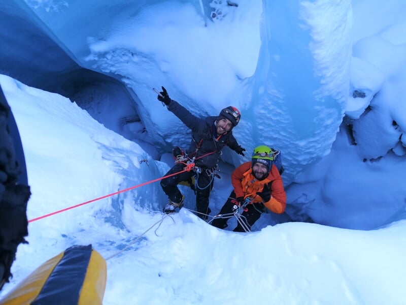 Deux spéléologues souriants équipés de cordes et casques dans une grotte de glace bleue.
