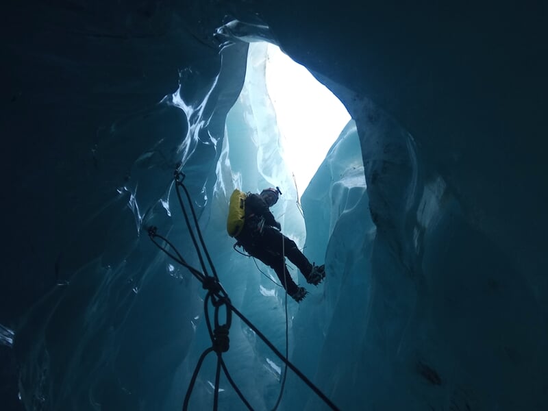 Spéléologue suspendu dans une crevasse glacée, entouré de parois de glace bleue lumineuse.