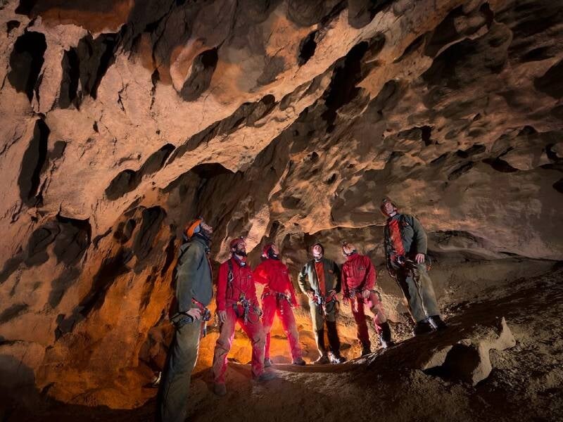 Groupe de spéléologues en combinaison rouge et casque dans une grande salle souterraine éclairée.