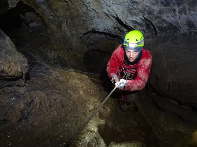 Spéléologue en casque vert descend en rappel dans une cavité rocheuse sombre et étroite.