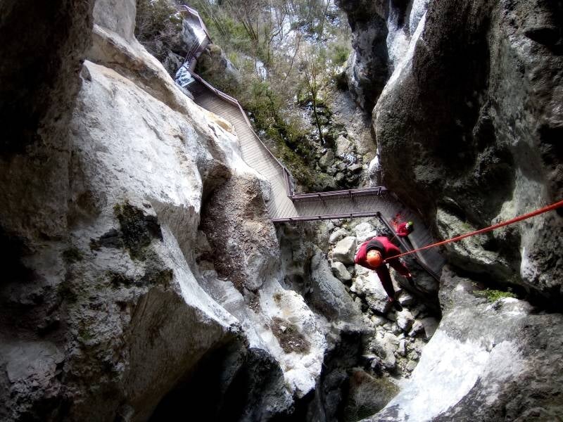 Spéléologue descend en rappel dans un passage rocheux étroit avec un pont métallique au-dessus.