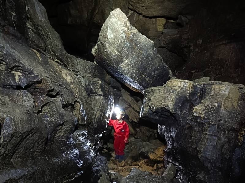 Spéléologue en combinaison rouge explore une grotte rocheuse sombre avec une lampe torche.