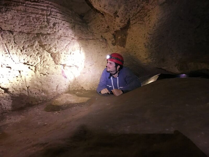 Homme avec casque rouge dans une grotte sombre, éclairée par une lampe, observant les formations rocheuses autour.