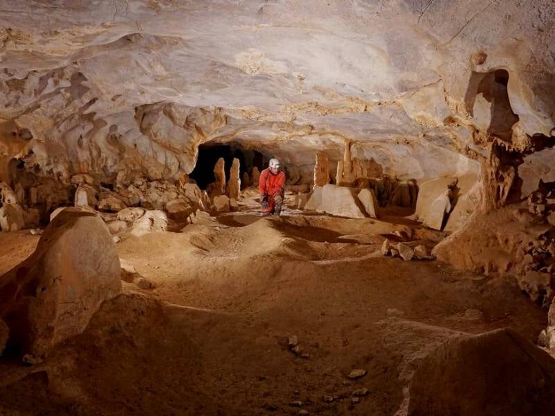 Explorateur en combinaison rouge dans une grotte avec formations rocheuses et sol sablonneux.