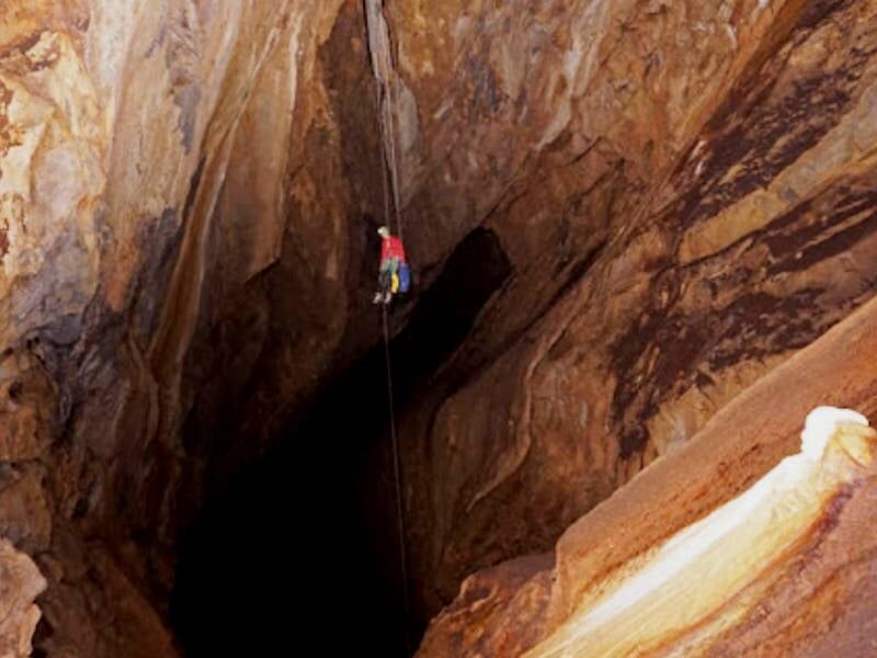 Personne en tenue de spéléologie descendant en rappel dans un puits profond et étroit de grotte.