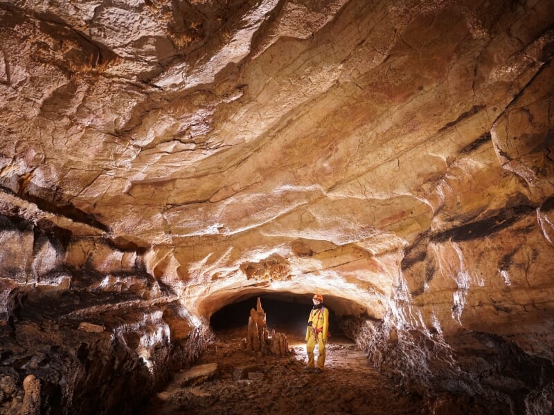Spéléologue en combinaison jaune marchant dans un tunnel souterrain aux parois lisses et éclairées.