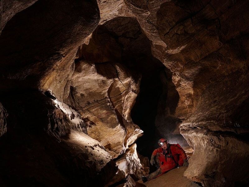Spéléologue équipé d'un casque et lampe explore une grotte rocheuse sombre aux parois irrégulières et étroites.