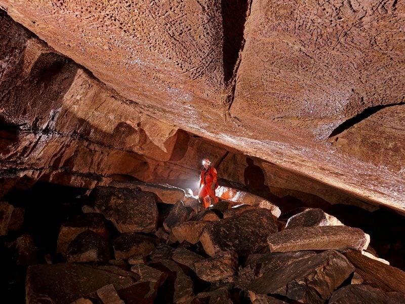 Spéléologue en combinaison rouge explorant une cavité rocheuse basse avec un casque éclairant les parois.