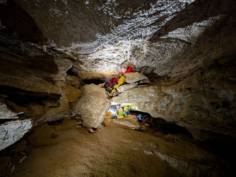 Spéléologues progressant dans une grotte avec un plafond haut, éclairés par des lampes dans un environnement rocheux.