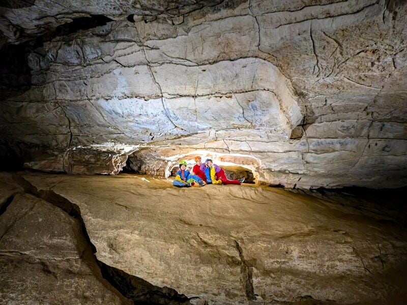 Quatre spéléologues assis sur une surface rocheuse lisse dans une grande cavité souterraine aux parois claires.