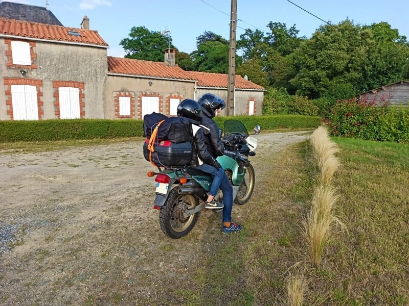 Motard en tenue noire sur une moto chargée, sur un chemin de terre près de maisons en pierre sous un ciel bleu.