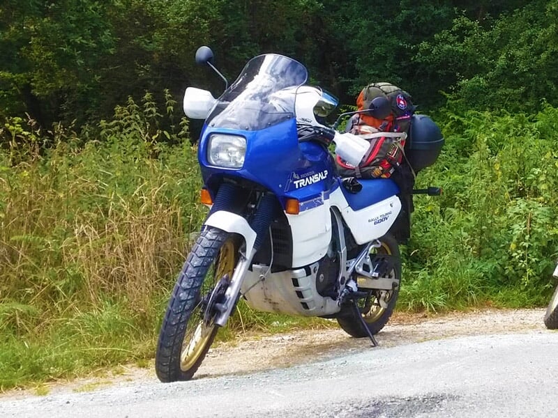 Moto blanche et bleue avec sacoches et casque, garée sur une route bordée de végétation dense et verte.