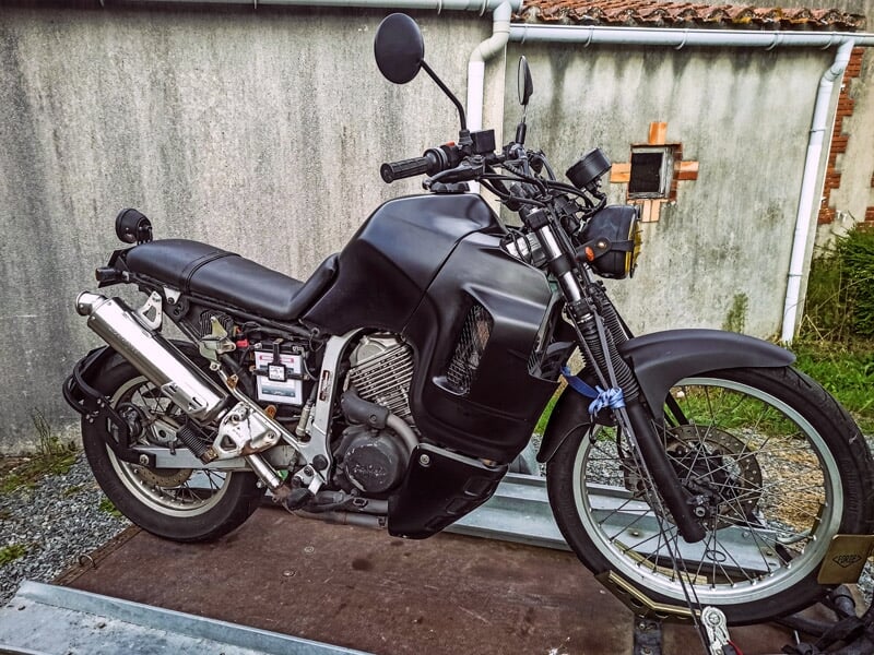 Moto noire avec cadre apparent, posée sur une surface en bois devant un mur gris, prête pour une balade.