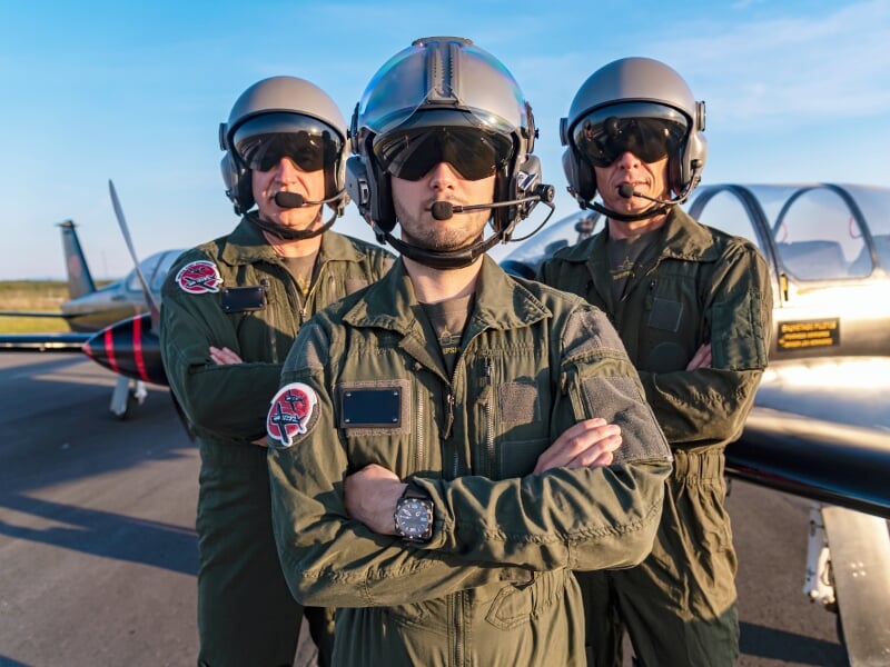 Trois pilotes en combinaison de vol debout devant un avion d'entraînement militaire sur le tarmac, avec des expressions confiantes