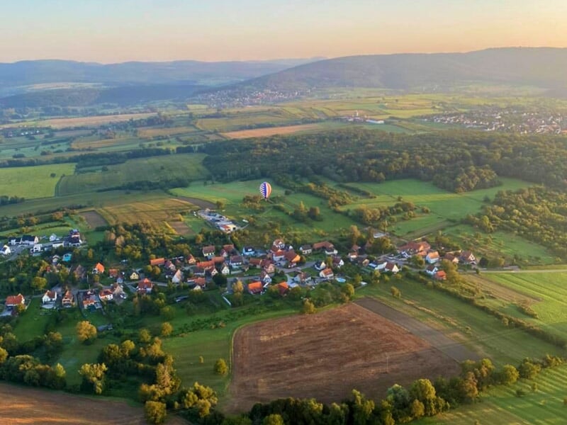 Vue aérienne d'un village entouré de champs verts et d'une montgolfière flottant au loin au coucher du soleil.