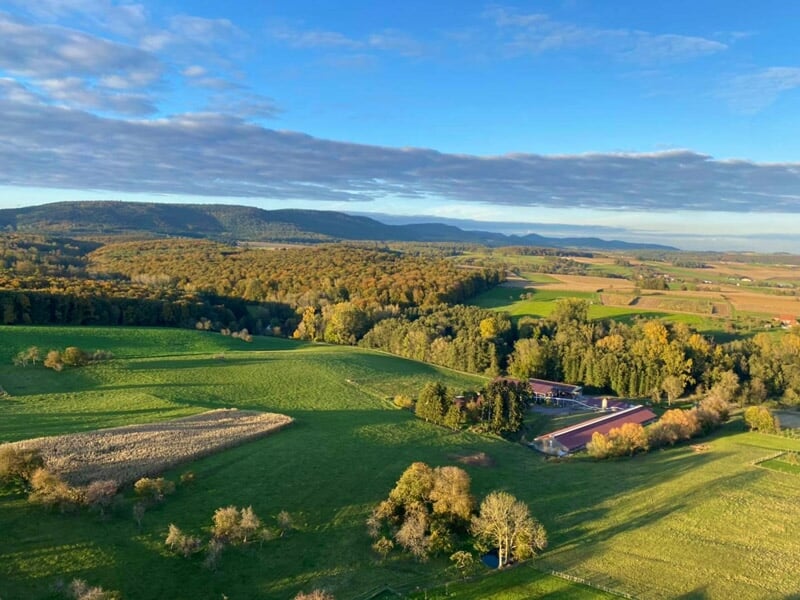 Paysage rural vu du ciel avec des champs verts, des arbres et une route, sous un ciel partiellement nuageux.