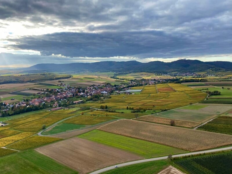 Vue panoramique aérienne de champs cultivés et d'un village au pied de collines sous un ciel nuageux.