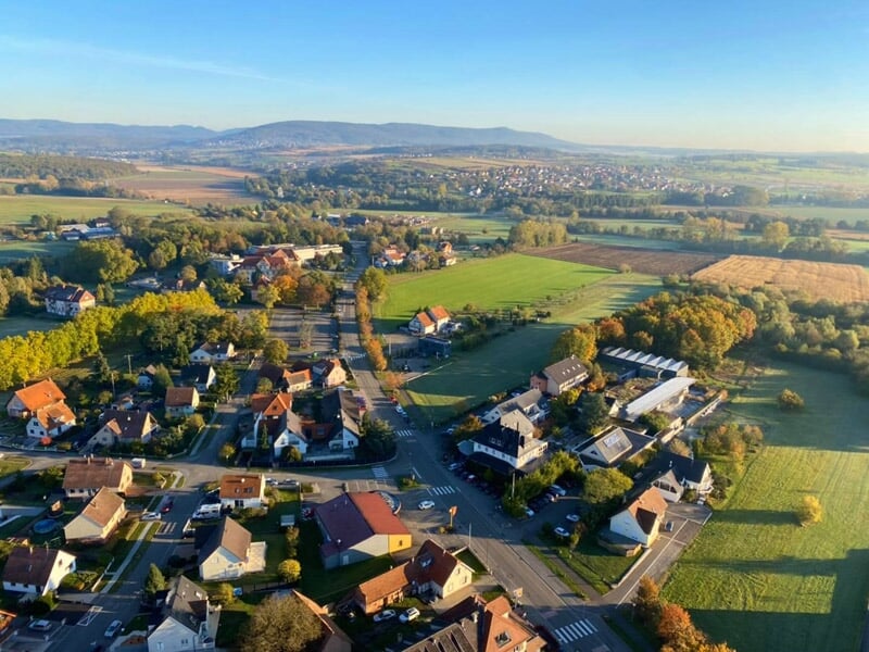 Village avec maisons et routes vu du ciel, entouré de champs verts et de collines à l'horizon au lever du jour.