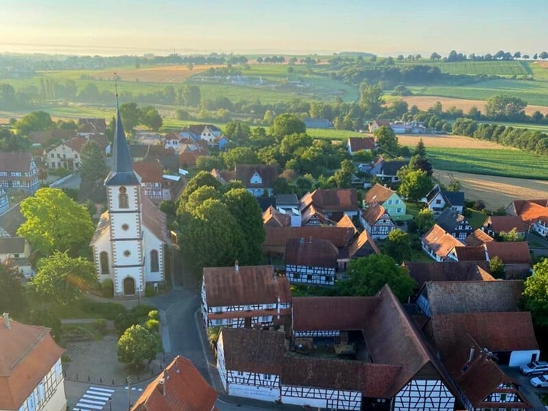 Vue aérienne rapprochée d'un village avec une église au centre, entourée de maisons et d'arbres verts.