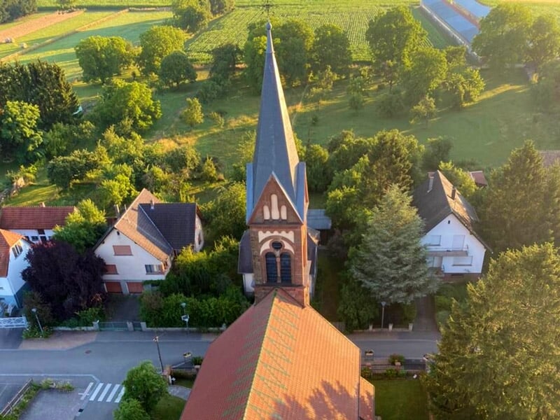Gros plan aérien sur une église avec un clocher pointu, entourée de maisons et de végétation ensoleillée.