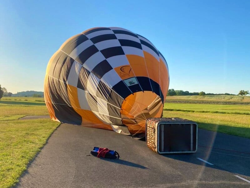 Montgolfière noire, blanche et orange dégonflée sur une route en campagne, avec un panier posé à côté.