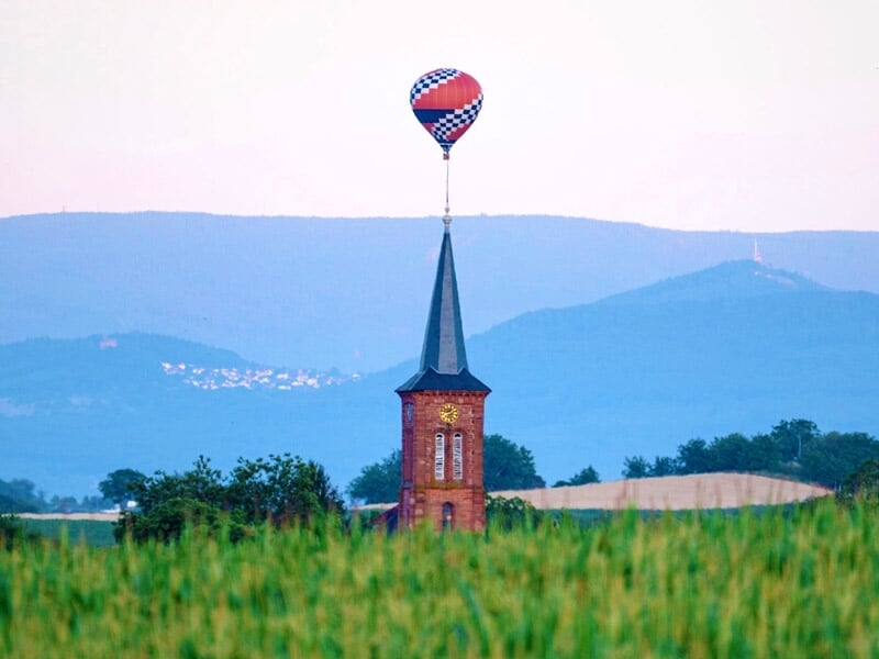 Montgolfière colorée flottant au-dessus d'un clocher d'église rouge dans un paysage de champs verts et collines.