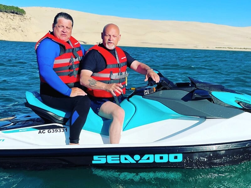 Deux hommes en gilets rouges sur un jetski Sea-Doo bleu et blanc avec une grande dune de sable en arrière-plan.