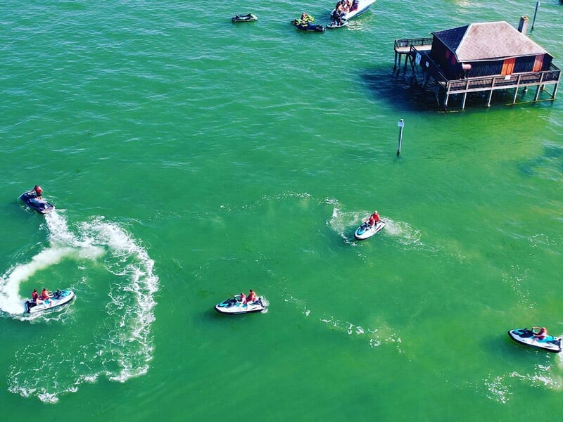 Vue aérienne de plusieurs jetskis circulant sur une eau verte près d'une cabane sur pilotis à Arcachon.