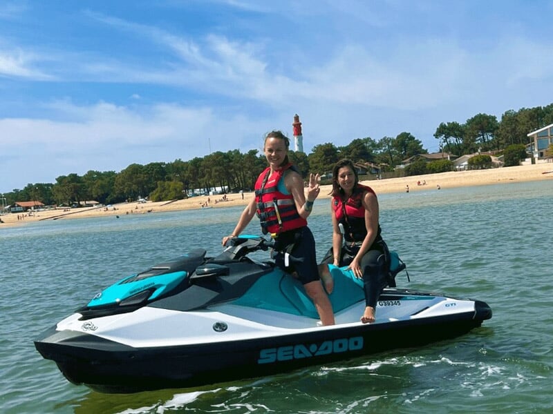 Deux femmes en gilets rouges posant sur un jetski Sea-Doo bleu et blanc avec une plage et un phare en arrière-plan.
