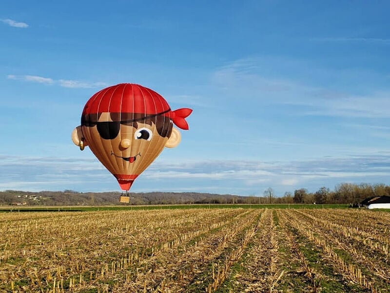 Montgolfière en forme de tête de pirate avec un bandeau rouge et un cache-œil, flottant au-dessus d'un champ cultivé sous un ciel dégagé.
