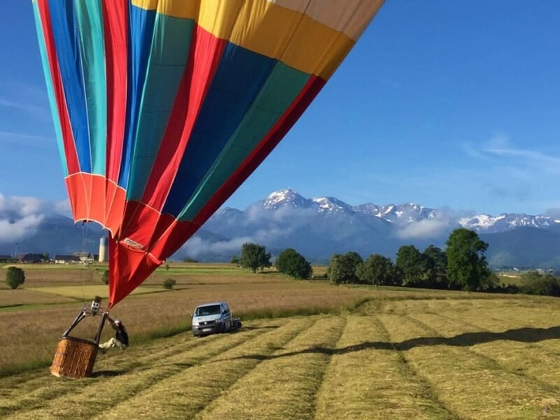 Montgolfière colorée au sol dans un champ fauché, avec les sommets enneigés des montagnes en arrière-plan sous un ciel bleu.