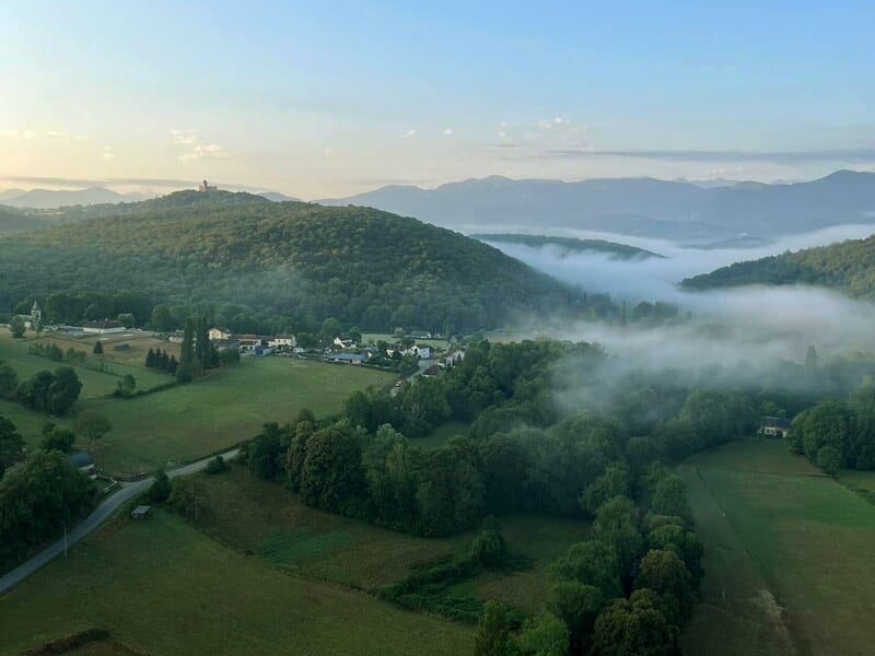 Vue panoramique sur une vallée verdoyante entourée de collines boisées avec des nappes de brume matinale, un village niché dans la campagne et des montagnes en arrière-plan.