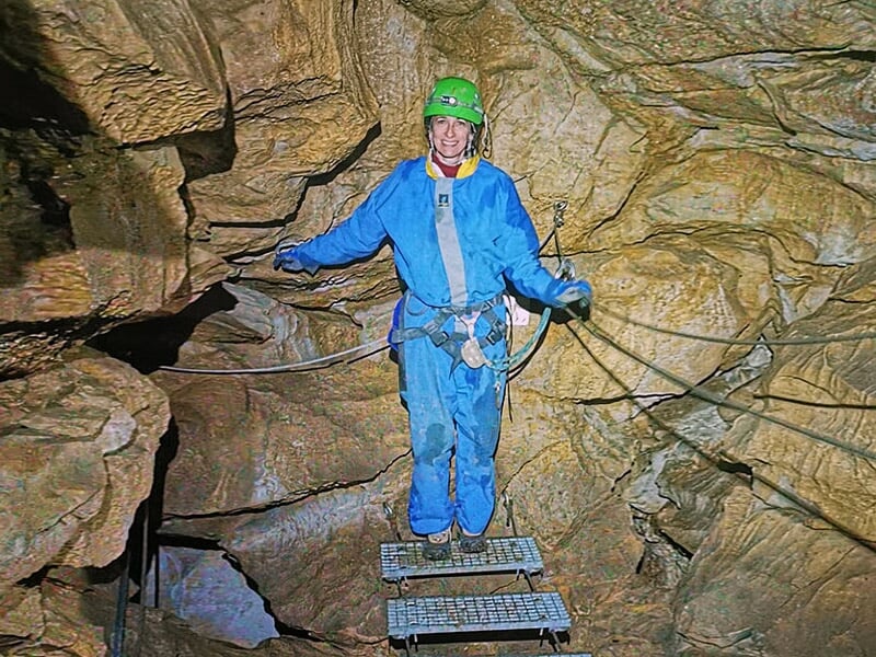 Spéléologue en combinaison bleue traversant un pont suspendu métallique dans une grotte.