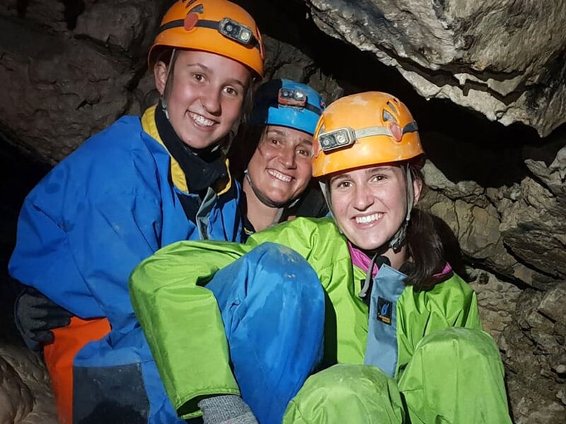Trois spéléologues souriants en combinaison colorée posant dans une cavité rocheuse souterraine.