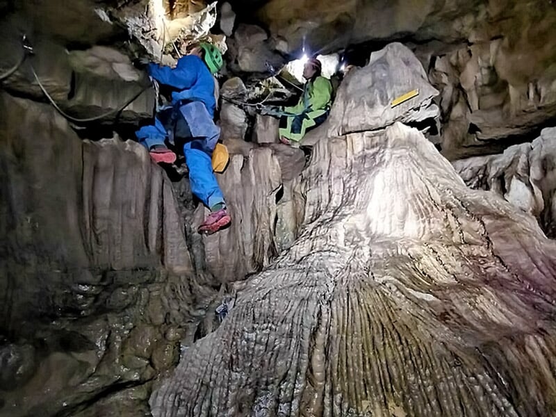 Deux spéléologues en combinaison bleue et verte escaladant une formation rocheuse dans une grotte.